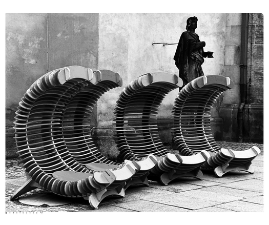 Aufnahme in schwarzweiß, vier hintereinander stehende Liegestühle, gebogen wie ein großes C (C wie Chemnitz) dahinter eine dunkle Statue aus Stein 