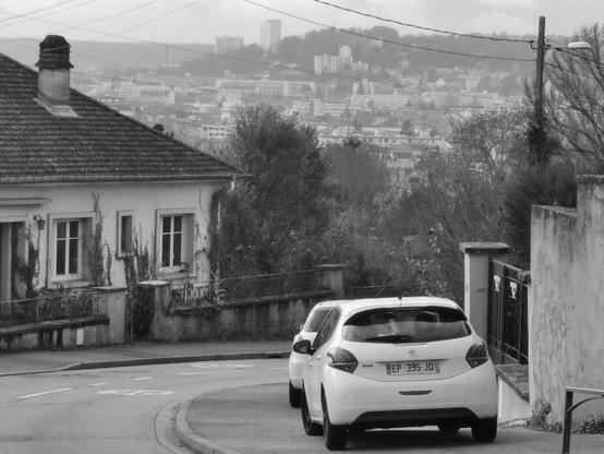 Vue d'une rue en pente en banlieue. Au loin le centre-ville apparaît en contrebas.