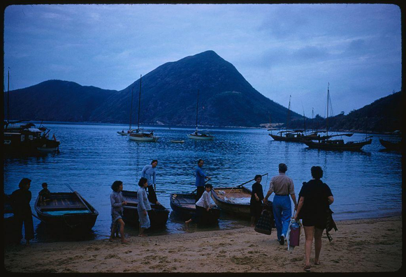 A group of people are gathered on a shoreline near boats and water. Some individuals appear to be boarding or disembarking from the vessels, while others walk along the shore carrying bags or engaged in conversation. The background features a scenic view with mountains rising against a twilight sky. There is no visible text within the image that provides additional context or information about its location or purpose.