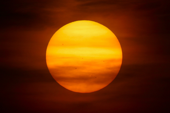 Photograph of a large and orange yellow sun against a dark orange sky with stripes from clouds and sunspots on the sun.