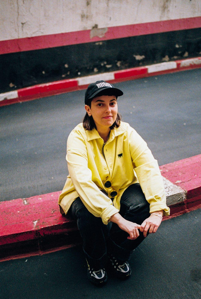 Fotografía de Andrea Galaxina sentada en la entrada de un garaje, vestida de amarillo y negro, rodeada de franjas negras, blancas y rojas.