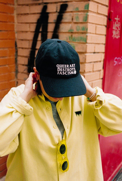 Fotografía de Andrea Galaxina. Tiene la cabeza inclinada hacia delante y viste una gorra donde puede leerse "Queer art destroys fascism".