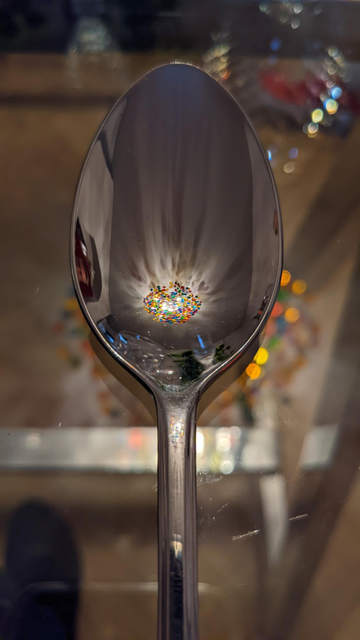 A mobile phone photo of a stainless steel spoon on a glass table. The deepest part of the bowl of the spoon has the reflection of an elaborate and decorative ceiling light. The latter composed of large glass beads in a wire matrix. Worth zooming in if you can.