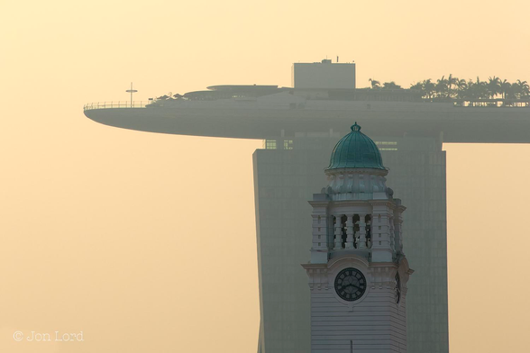 This is a colour sunrise photo in landscape format of a colonial era historic clock tower with contempory high-rise building in the background. Singapore (2015).

Below and to the right of centre is a square, white clock tower with a round dial clock in each corner, with two clock faces in view: one facing the camera and one viewed side-on. The clock face is white with the time in black Roman numerals, the clock hands also in black. The time indicated is nineteen minutes past eight. Above are square colums with a few bells visible inside. On top is a copper-green domed roof. In the background and immediatly behind, perhaps one km away, is the tall, rectangular tower of a high-rise hotel. On top of the high-rise is a large arched and slightly surfboard shaped deck that stretches from beyond the tower to the left, over the tower and off the photo to the right. The top of the deck has railings around the edge, made to look minature by the size of the deck. There are some one story structures and further to the right numerous small palm trees. Surrounding this scene and in the background is a dull orange sky, coloured by the low morning sun and probably atmospheric pollution.

The location: Marina Bay, Singapore. The clock tower belongs to the Victoria Theatre and the high-rise is the Marina Bay Sands hotel, casino and shopping centre.
