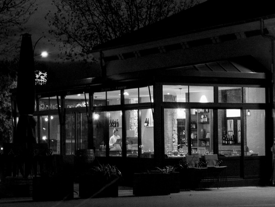 Une photo en noir et blanc, de nuit, format paysage, qui montre la structure légère d'un restaurant aux grandes baies vitrées éclairées de l'intérieur.  Une jeune femme lit ce qui ressemble à la carte du restaurant en attendant, peut-être, de prendre son service au milieu des tables dressées. Sur la placette devant le bâtiment  quelques tables et chaises de grandes jardinières fleuries, et sue la gauche, la silhouette d'un parasol replié.