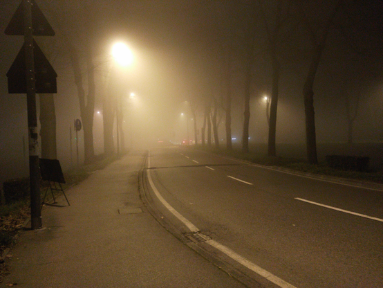 A foggy street at night with dim streetlights casting a warm glow, bare trees lining the road, and the sidewalk fading into thick mist while distant headlights appear blurred.