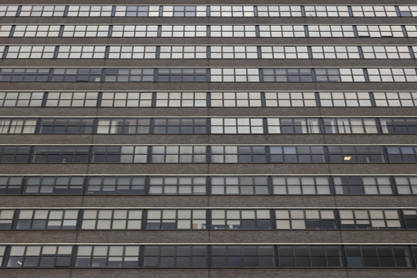 A close-up view of a modern building’s façade, showing multiple rows of rectangular windows set in dark frames. The walls between the windows are made of brown brick, creating a striped pattern with the alternating glass panes.