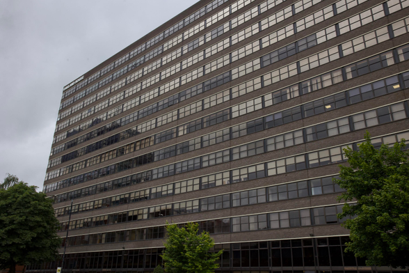 A large, multi-storey office building with many rows of windows and a brick façade, viewed from a low angle. Trees with green leaves are in the foreground, and the sky above is overcast and grey.