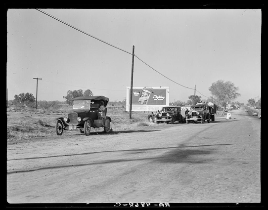 The image is a black and white photograph depicting an outdoor scene from the early 20th century. It shows three vintage cars parked on the side of a rural road, with two billboards visible in the background. One billboard displays what appears to be an advertisement for Coca-Cola Oil Film, while the other seems obscured or less clear due to the angle and distance.

The cars are old-fashioned models from that era; they have large bodies, prominent grilles, and spoked wheels typical of early automobile design. The car on the far left is closest to the viewer with its front license plate visible. In between these two vehicles stands another older model, which appears more compact compared to the first one.

The road itself has a simple, unpaved surface that suggests it's in a rural or less developed area. A telephone pole runs parallel to the roadway on the left side of the photo, adding to the rustic setting. The background is relatively barren with minimal vegetation and structures; however, some trees can be seen at various distances.

There are no people discernible within this particular frame that would suggest their occupation beyond driving vintage cars. No specific context or story about "Three carloads of Mexicans headed for the Imperial Valley" appears directly in the photograph but it is referenced as a source link to obtain more information on Dorothea Lange's  [...]