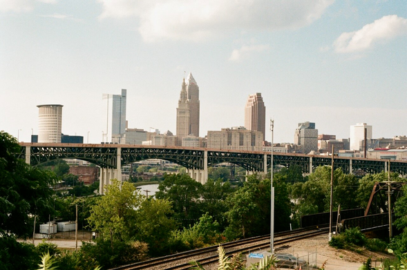 In the foreground, railroad tracks cross the lower scene leading to a bridge. The area is heavy with trees. Hope Memorial Bridge breaks the scene up in the center dividing the fore and back grounds, with some old brick buildings tucked below the bridge to the left. In the background, the Cleveland Skyline shows above the bridge on a nice summer evening.