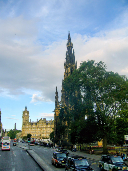 A high-contrast 2006 photograph taken on a city street in Edinburgh, Scotland, under a dramatic sky of patchy blue and white clouds. The main subject is the Scott Monument, a massive, dark, Victorian Gothic spire that takes up the right side of the image, appearing rugged and weathered. Its intricate, multi-tiered structure reaches high into the frame. In the mid-ground to the left, standing slightly shorter, is the handsome, buff-colored stone building of the Balmoral Hotel, featuring a prominent clock tower. The street below is busy, with several traditional black London-style taxis and one red double-decker bus moving away from the viewer. A row of dark-green fencing separates the monuments and a large, full-leafed tree from the paved road, where pedestrians are walking on the footpath to the right.