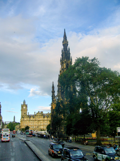 A high-contrast 2006 photograph taken on a city street in Edinburgh, Scotland, under a dramatic sky of patchy blue and white clouds. The main subject is the Scott Monument, a massive, dark, Victorian Gothic spire that takes up the right side of the image, appearing rugged and weathered. Its intricate, multi-tiered structure reaches high into the frame. In the mid-ground to the left, standing slightly shorter, is the handsome, buff-colored stone building of the Balmoral Hotel, featuring a prominent clock tower. The street below is busy, with several traditional black London-style taxis and one red double-decker bus moving away from the viewer. A row of dark-green fencing separates the monuments and a large, full-leafed tree from the paved road, where pedestrians are walking on the footpath to the right.