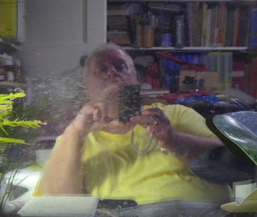 Selfie of a fat man in a yellow t-shirt reflected in a dirty window.