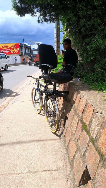 A person sits on a brick wall beside a bicycle, which has a black swivel office chair strapped to its frame. The scene is set on a sidewalk next to a busy street in Antananarivo, Madagascar, with a billboard and passing vehicles visible in the background.