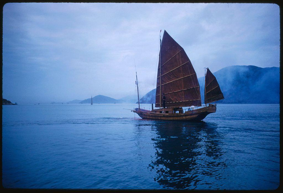 A large sailing ship with two tall, brown sails is gliding across a calm body of water under overcast skies. The background shows distant mountains partially obscured by mist or low-lying clouds. Another small boat can be seen in the far distance on the left side of the photo. This image appears to capture a serene moment from a sightseeing tour in Hong Kong, as documented through a photograph taken during July 1959 and presented here with detailed context provided for visual description purposes.