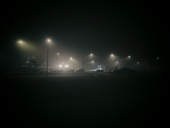 A foggy parking lot illuminated by streetlights, with several cars visible in the foreground and background. The atmosphere is dim and mysterious due to low visibility.
