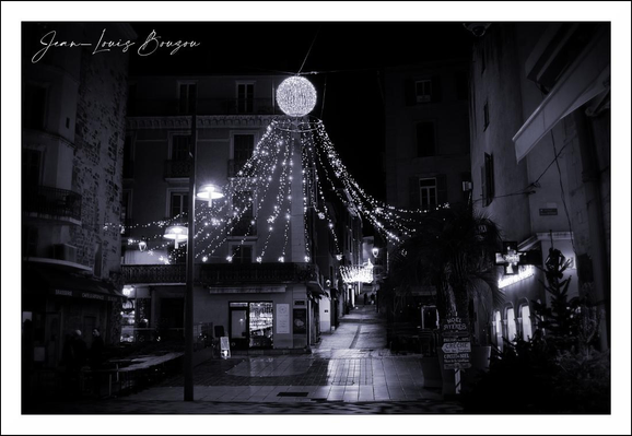 A quiet, nocturnal street scene rendered in high-contrast black and white. At the center a bright, spherical lamp hangs above the lane, and from it dozens of small lights cascade outward like a festive canopy or a constellation poured down over the buildings. The strands form a starburst that draws the eye to the center and then down along the wet cobblestones, which mirror the glow in soft, fractured reflections.
The narrow street is framed by low buildings with shuttered windows and small shopfronts; their facades recede into shadow, giving the scene a compressed, intimate feeling. A few wall-mounted lamps and illuminated signs pick out architectural details — a balcony railing, a doorway, the texture of stucco — while most of the façades sink into velvety darkness. On the right, a cluster of foliage or a potted tree interrupts the geometry, its silhouette softened by the lights.
There is a stillness to the composition: no moving cars or busy crowds, only the gentle play between light and shadow and the suggestion of human presence in the open doors and lit windows. The overall mood is at once celebratory and contemplative — a small, luminous celebration suspended above a quiet, sleeping street.
