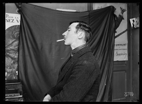 A man is seen standing with his head slightly tilted to the side, wearing a dark jacket and smoking a cigarette. He appears contemplative or possibly in deep thought as he gazes off into the distance. The setting seems informal, perhaps an indoor environment like a studio or workshop. There are various objects around him including what looks like a poster on the wall with text that is partially legible: "Pour la Sez V...". Another sign reads "Sauvez," which translates to “Save” in English. A door labeled "3740" and another one stating "sauveur" or "rescuer", are also visible, suggesting a possible connection with military or rescue services during wartime. The overall tone is reminiscent of historical photos capturing significant moments from the past, possibly related to World War I given references like 'Sauvez' which could be associated with war-time efforts and support for soldiers.