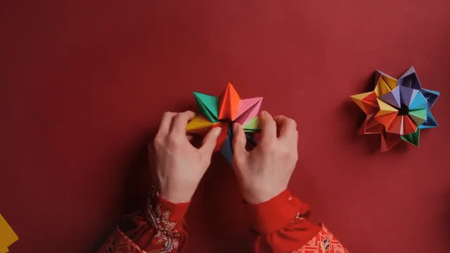 Hands manipulate a colorful paper origami kaleidoscope toy against a red background, with a completed multicolored kaleidoscope visible to the right.
