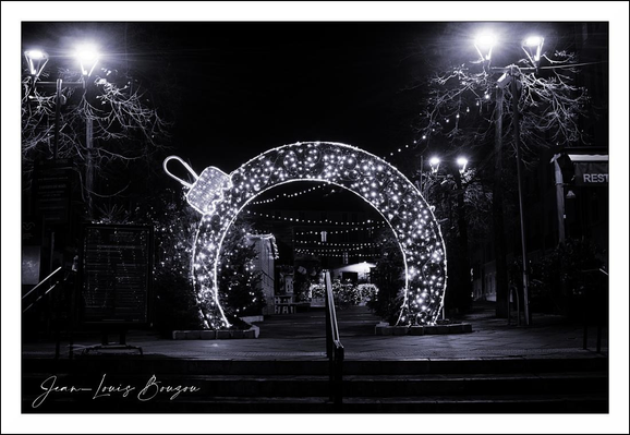 A quiet urban night, rendered in stark black and white. At the center of the frame a giant ornament-shaped arch, studded with countless tiny lights, forms an illuminated portal — a luminous circle with the loop of a bauble perched at the top — and invites you to pass through its glow.
String lights trail away through the arch, drawing the eye down a darkened path and giving the scene a gentle sense of depth and repetition. The light from the installation pops against the surrounding darkness, creating a high-contrast tableau where every bright filament feels like a star against velvet night.
Two streetlamps stand to either side, their halos flaring and catching the bare branches of the trees, which reach in like skeletal fingers. The trees and lamps frame the composition, adding vertical counterpoints to the arch’s roundness and reinforcing the symmetry of the scene.
In the foreground, steps descend toward the ornament; a handrail bisects the lower part of the image, grounding the viewer and suggesting a threshold between the ordinary street and this pocket of celebration. The pavement reflects a touch of light, hinting at recent rain or the sheen of cold air.
A lone silhouette moves beneath the lights, small and softly defined — a human presence that lends scale and a quiet, contemplative loneliness to what would otherwise be pure spectacle. There’s a stillness to the photograph, an almost cinematic pause between moments of movement.
