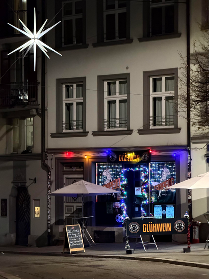 Eine nächtliche Szene mit einer gemütlichen Bar, die mit festlichen Lichtern und einem großen Stern darüber geschmückt ist. Die Bar mit dem Namen „torpedo“ zeigt Schilder, die auf Getränke hinweisen, und verfügt über Sitzgelegenheiten im Freien unter Sonnenschirmen. Das Gebäude hat mehrere Fenster, und die Atmosphäre deutet auf einen lebhaften Urlaub hin