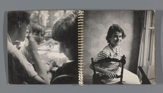 The image depicts a man with curly hair wearing glasses, sitting and looking down at what appears to be an object in his hands. He is surrounded by other people who seem engaged in conversation or observing something off-camera. The setting looks casual and social, possibly outdoors during daytime hours as indicated by the natural light illuminating their faces.

The photograph has a vintage feel with its black-and-white composition and classic attire worn by the subjects. It evokes a sense of nostalgia for past times when such candid interactions were more common in everyday life.