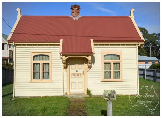 A centred shot of a historic, single story wooden building with light yellow horizontal siding and a reddish brown corrugated iron roof. The door is labelled "POLICE." Two arched windows flank the central entrance.