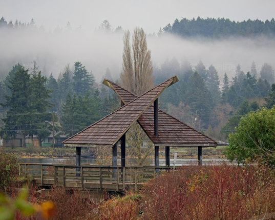 A shelter further along the path is seen with its distinctive two roofs that cross at an angle. A tall tree in the background is centred at their intersection. The hills in the far background sport low lying cloud that nearly touches the sea