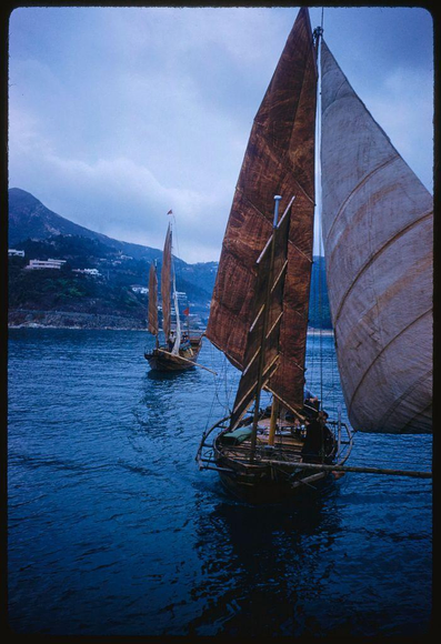 Two sailboats with brown and white sails on a body of water, likely in Hong Kong given the collection's reference. The boats have wooden structures supporting their masts and sails, suggesting they are traditional or recreational vessels rather than modern racing yachts. There is visible movement in the ripples across the blue waters under an overcast sky. In the background, mountains can be seen with a few buildings on them, hinting at a coastal urban area. The overall atmosphere of this photograph suggests it may have been taken during a cooler or cloudy day, as indicated by the muted colors and lack of direct sunlight reflection off the water's surface.