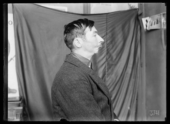 The image is a black and white photograph, likely taken in the early 20th century given its style. It features a man standing profile to the camera against an indoor backdrop that appears informal; it includes draped curtains or drapes on either side of him with various indistinct objects scattered above them, such as what might be papers affixed to boards. The lighting is flat and suggests artificial light typical for studio settings.

The man has short hair combed back from his forehead, a full face sporting stubble that's not entirely shaved or neatly groomed, which was common in working-class men during this period. His expression seems neutral but attentive; he appears contemplative rather than smiling or frowning directly at the camera. He wears what looks like an early 20th-century style coat with a collar and button-down shirt underneath.

There's also text '3741' on the lower right side of the photo, possibly indicating its catalog number within a larger collection, suggesting that this image was part of an archival or documentary series. The man’s posture is relaxed yet somewhat alert as if he were awaiting something or someone during his brief moment in front of the camera.

The photograph's overall composition and setting suggest it could be from Lewis Hine's work documenting various aspects of American life at the time, including laborers' conditions which ma [...]