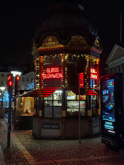 An old fashioned kiosk at a Copenhagen Christmas market late at night. The stalls are all closed, the kiosk too, but the lights are still on. Red neon signs announcing GLØGG and GLÜHWEIN. Inside the kiosk, lit up in white light, one can make out the wine glasses, hanging upside down from their rack, and paper cups, for coffee. To the left a red traffic light, and in the background trees lit up in gold and blue.