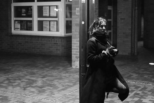 B&W-Nachtaufnahme einer jungen Frau. Sie steht - in einer Fußgängerzone- an einen Laternenmast gelehnt. 