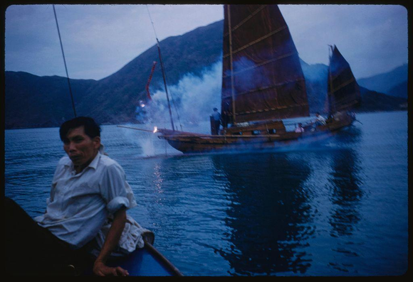 This photograph features a man on the left side of the frame, sitting in what appears to be an open boat. He is wearing a light-colored shirt and has dark hair. The background shows another large sailboat with its sails unfurled moving through the water, creating waves behind it as well as some smoke or steam coming off from somewhere near the back part of the vessel. There are several people visible on this second boat; however, their features aren't clear enough to discern any specific actions they might be engaged in.

The setting appears to take place during twilight hours with a soft blue hue lighting up both boats and reflecting off the water's surface. The surrounding landscape includes mountainous terrain that adds depth to the scene. Overall, it gives an impression of a serene but active maritime environment captured through color transparency film on 35 mm slide format.

Additional context suggests this photo was taken by American photographer Toni Frissell during her assignment for Sports Illustrated in Hong Kong (Job Number: x5826). It seems to depict sightseeing activities within the city, reflecting both cultural aspects and daily life experiences.
