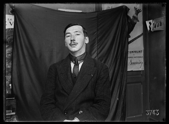 The image is a black and white photograph, likely from the early 20th century given its style. It depicts an individual sitting in front of what appears to be a backdrop with text, suggesting they may have been photographed for identification or military records during that era.

The person has a mustache, wears glasses held up by wire rims, is dressed formally in dark clothing including a jacket and tie. The attire indicates he might hold some level of authority or rank within the army as suggested by his uniform-like appearance with epaulettes on each shoulder which are typical features for military uniforms.

The backdrop includes text that seems to be related to French language signs ('la v', 'l' emprunt'), possibly indicating this photo is connected to a specific event, location (such as France), or unit within the army. There's also a number at the bottom right of the image - "3745" which could indicate an identification code for records.

The overall condition and style suggest that it may have been taken in Paris given its language context.