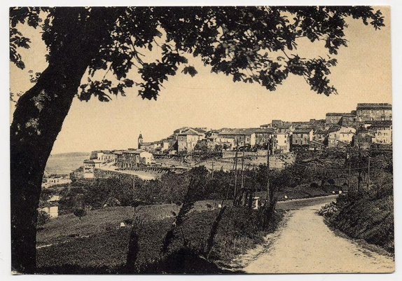 This black and white photograph shows a panoramic view of Città della Pieve, with the town's architecture prominently featured. The perspective is from an elevated vantage point overlooking the village nestled amidst rolling hills or terraced vineyards. A prominent tree branch on the left frames part of the image, drawing attention to the top edge where its silhouette contrasts against a lighter sky background.

The buildings are predominantly two and three stories tall with pitched roofs typical of Italian architecture. The central focus is likely a church steeple, indicating religious significance within town's layout. There’s a pathway leading towards one side of the village that suggests accessibility for visitors or residents alike. Surrounding greenery indicates agricultural land use below.

The image has an antique feel to it, suggesting historical context and possibly capturing life in Città della Pieve during early 20th-century times as indicated by its style and quality. It's worth noting there are no visible people which can make the scene appear tranquil or unoccupied at this moment captured within time.

The photograph is part of a collection associated with Ferro Candilera, whose link suggests it may be part of an exhibition or historical record related to his works on Italy's landscapes and architecture.