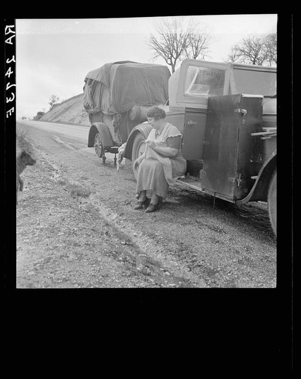 The image depicts a black-and-white photograph of a woman seated on the running board of an old-fashioned truck. The vehicle is heavily laden with what appears to be household items, including furniture covered in dark tarps and other personal belongings stored within crates or cases attached to the sides of the trailer hitched behind it. This suggests that they are migrant workers traveling with their possessions during a journey.

The woman seated on the running board wears a modest dress and has her hands clasped together, possibly indicating fatigue or contemplation. Her gaze is directed off-camera, adding an element of mystery as we do not know where she might be looking at in this moment captured by Dorothea Lange's camera lens.

In the background, there are leafless trees, which could indicate a colder season when many migrants travel to find work during harvest seasons or warmer months. A dirt road stretches into the distance under an overcast sky, emphasizing the hardship and journey these individuals endure in their quest for better living conditions and employment opportunities away from home.

This image is part of Dorothea Lange's documentation on migrant workers, as indicated by the caption mentioning "Migrant pea workers on the road." The photograph offers a poignant glimpse into the lives of those displaced due to seasonal labor demands.