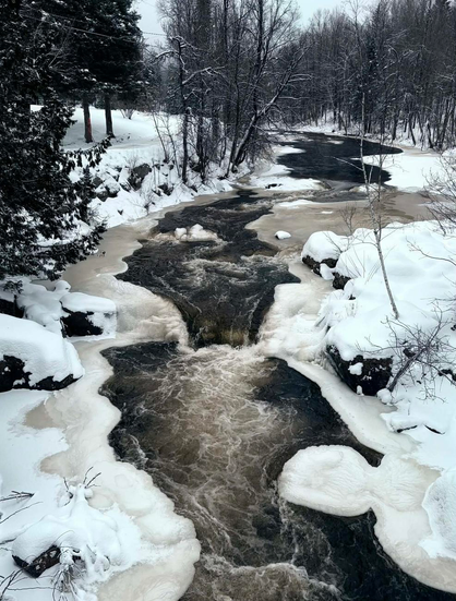 Photograph of a river partially covered in ice and snow, with a fast current, a small waterfall and rapids, bordered by forest on both sides. The photo is taken from above, from a bridge over the river.

Photographie d'une rivière partiellement recouverte de glace et de neige, avec un débit rapide, une petite chute et des rapides, bordée de forêt de chaque côté. La photo est prise de haut, à partir d'un pont au-dessus de la rivière.