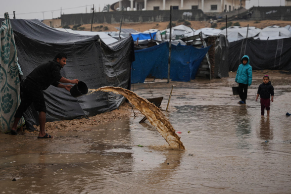 A man clears water from his tent at a camp for displaced Palestinians after heavy rain, as children watch on.
