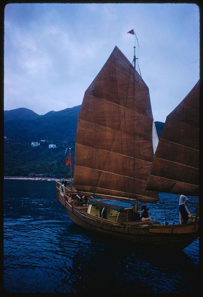 A traditional sailing vessel with large brown sails is seen on a body of water surrounded by hills and a partly cloudy sky. The ship has two visible masts, each adorned with several square sails made from an earthy-colored fabric. Flags are attached to the topmost mast, adding detail to this distinctive sailboat design. Onboard, there appear to be at least four people: one in white standing near the back of the boat under a shaded area, another wearing pink and black sitting on the deck closer to the stern, two more individuals partially visible; they are dressed casually with hats or head coverings.

The scene captures both human activity and natural beauty. The water is calm, reflecting light from various sources including what might be early evening sunlight that casts a warm hue over parts of the image. In the background, buildings on a hill suggest an urban environment not far away from this serene nautical setting. Overall, it's a picturesque moment captured in time by Toni Frissell during her assignment for Sports Illustrated.

Note: The date mentioned in your prompt is July 1959 which could be relevant to understanding the historical context of Hong Kong at that time as well as providing information about when this photograph was taken or intended.