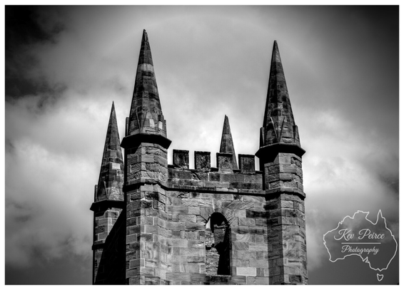 Black and white, low angle photograph of the stone ruins of the Port Arthur Penitentiary Church tower in Tasmania.  The tower features battlements and four distinct stone spires pointing skyward. A subtle but clear solar or cloud halo arcs over the central, tallest spires.  The texture of the old, weathered stonework is highly detailed and contrasts sharply with the dramatic, cloudy sky.