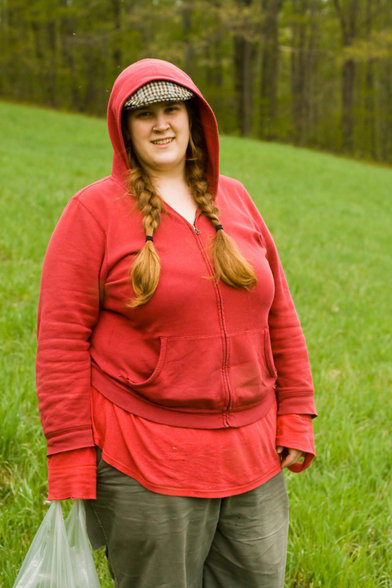 A woman wearing a red hoodie and a checked hat, grey pants and twinbraids is holding a plastic bag as she stands on a slanted grassy field with a hedgerow in the background