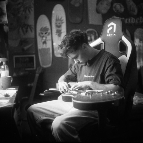 Black and white photo of a young man sitting in a room, focused on adjusting a guitar on his lap, surrounded by skateboards and personal objects, quiet and intimate atmosphere.