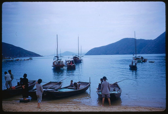 A group of people and boats at the shore, with mountains in the background. The image depicts a serene scene on calm water near Hong Kong during what appears to be sunset or dusk. Several individuals are seen interacting around small wooden fishing vessels while others row further into the distance using oars. Some wear hats, jackets, and casual clothing suitable for outdoor activities. A few boats have masts standing erect against the sky. The color palette is dominated by soft blues of the water contrasted with muted earth tones on land. No specific event or occasion seems to be taking place; rather it captures a slice of daily life near Hong Kong's waterfront.