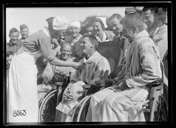 The image is a black and white photograph depicting an interaction between healthcare workers and patients in wheelchairs. The focus of the photo captures two individuals, both seated on what appear to be manual wheelchairs with spoked wheels typical of early or mid-20th-century design. One individual is wearing striped pajamas commonly associated with hospital attire for convalescent care.

The person standing appears to be a healthcare worker, possibly female due to the long apron and head covering reminiscent of traditional nurse's clothing from historical periods. The caregiver extends an arm towards one of the seated individuals in a gesture that suggests offering assistance or interaction.

In the background, several other figures can be seen smiling and observing the exchange between the standing person and the individual on the wheelchair closest to them. These individuals are also dressed informally, some with casual clothing like trousers rolled up, which may indicate they too have been patients but now in a more relaxed state of recovery or convalescence.

The setting is outdoors during daylight hours as indicated by the shadows cast on the ground and bright ambient lighting reflecting off surfaces within the scene. The photograph has an alphanumeric code "8063" handwritten at the bottom left corner, likely for cataloging purposes. This image evokes a sense of  [...]