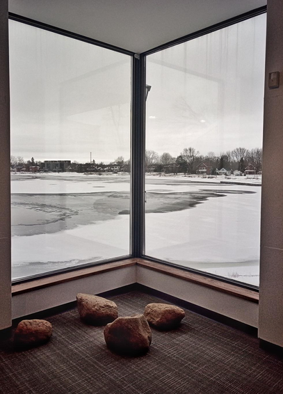 Looking out of a large two paned corner glass window onto a partially frozen lake. Snow and ice can be seen covering some of the water and trees and houses are shown in the background. Four large rocks sit on the carpeted floor of the interior of the building. 