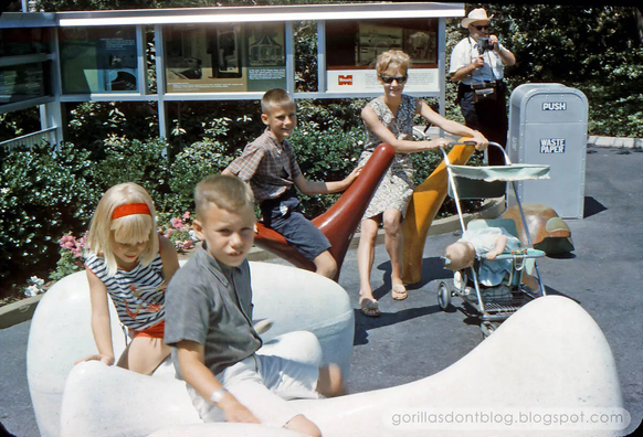 Vintage theme park scene with children sitting on sculpted seats shaped like abstract curves, an adult pushing a stroller, and a “Waste Paper” trash can nearby. A display board with photos and text stands in the background under sunny skies.