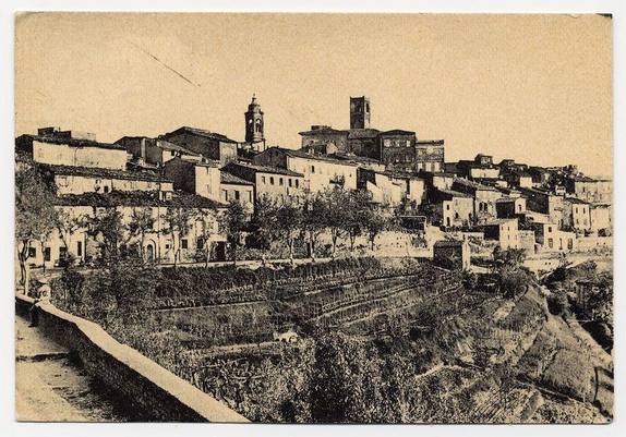The black and white image displays a historic town perched on elevated terrain, likely captured in the early 20th century. The settlement features clustered buildings with varying rooflines, indicative of different architectural styles, some displaying signs of historical wear or age. Prominent structures include what appears to be a church tower and another tall structure that could serve as an observation point.

The town is surrounded by terraced fields suggestive of agricultural activity, possibly vineyards given the regularity and arrangement typical for wine production regions in Europe. A stone wall runs along one edge of these fields, with sparse vegetation scattered throughout what appears to be a dry or temperate climate landscape. There's no visible modern technology or infrastructure that would place this image more recent than historical.

The overall composition gives an impression of a tranquil and timeless rural setting, possibly capturing the everyday life in Città della Pieve during its earlier days. The absence of people adds a sense of stillness to the scene, inviting viewers to ponder on what daily life was like here long ago.