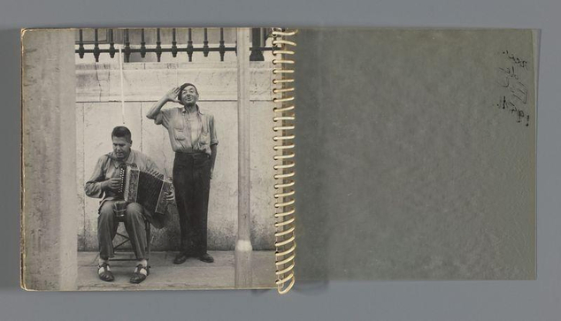 The image is a black and white photograph displayed in an open spiral-bound notebook or album. On the left page, there are two men standing outside against a backdrop of a building with barred windows and metal railings on top. The man seated to the left appears focused on playing an accordion while holding another one under his arm; he wears casual clothing consisting of trousers, shoes, and sandals, and has glasses perched atop his head. Beside him stands the second man in what seems like a defensive or protective stance with his hand raised to his forehead. He also dresses casually but adds a pocket watch chain around his neck.

On the right page is an open gray notebook, seemingly unmarked aside from some faint text near its top edge which appears handwritten and possibly of foreign language script. The overall impression suggests this might be part of a photo album documenting street musicians or everyday life scenes captured by photographer Ed van der Elsken during travels in Marseille between 1948-50.

The image conveys an air of candid urban photography, capturing moments from the lives and surroundings of individuals on the streets.