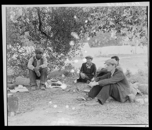 The image is a black and white photograph showing three men seated on the ground in an outdoor setting. They appear to be engaged in conversation, with one man looking at another while he gestures or speaks. The background reveals sparse vegetation and what seems like remnants of temporary structures, suggesting this could be a work campsite.

Each man is dressed differently but generally wears casual attire appropriate for manual labor: hats, jackets, and trousers. One has his hands clasped in front of him; others have their arms resting on the ground or holding objects that are not clearly visible due to resolution limits. Various items such as cups, a mug, containers, and possibly utensils lie around them.

The photograph is marked with text "IN DORE BE AR," which could indicate a specific location or context within Nipomo, California where pea fields might be located near the roadside mentioned in the caption. The overall impression suggests an historical snapshot of workers taking a break during fieldwork activities, possibly from agriculture given the title reference to peas and roadsides.

This image is labeled as "Camp of single men by the roadside" with additional information provided about it being captured in Nipomo, California, which aligns with the visual cues within the photograph.