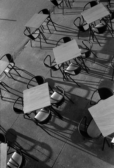 An overhead view of a number of wooden tables and black chairs arranged on a concrete surface, casting shadows in black and white.